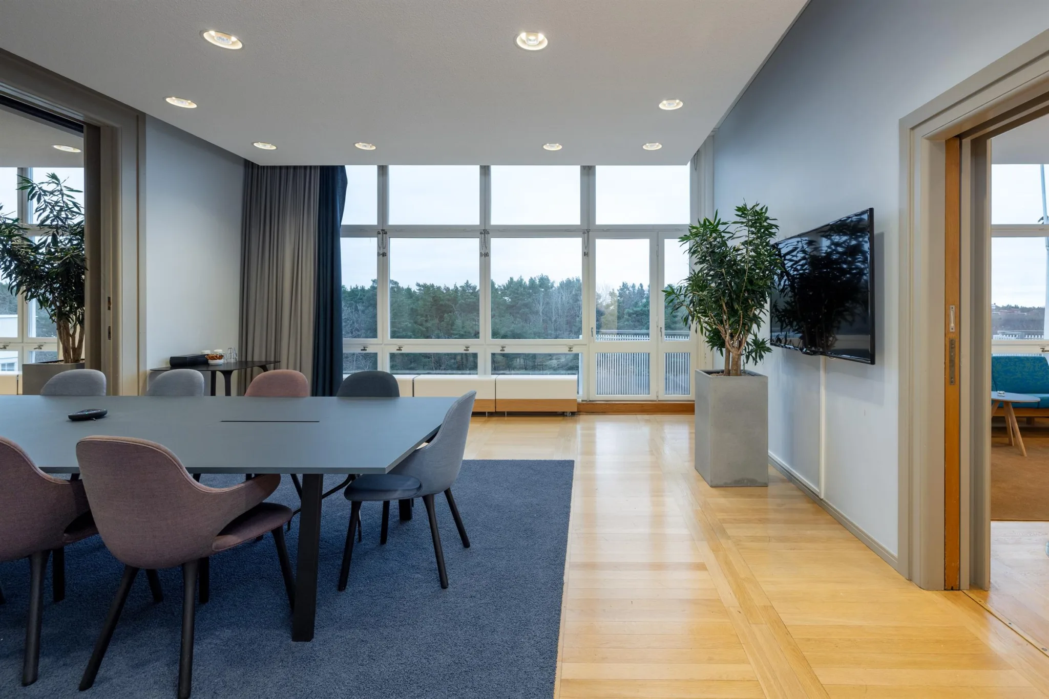 3. Ett konferensrum med ett bord, stolar, en stor TV på väggen och höga fönster med utsikt över grönska.