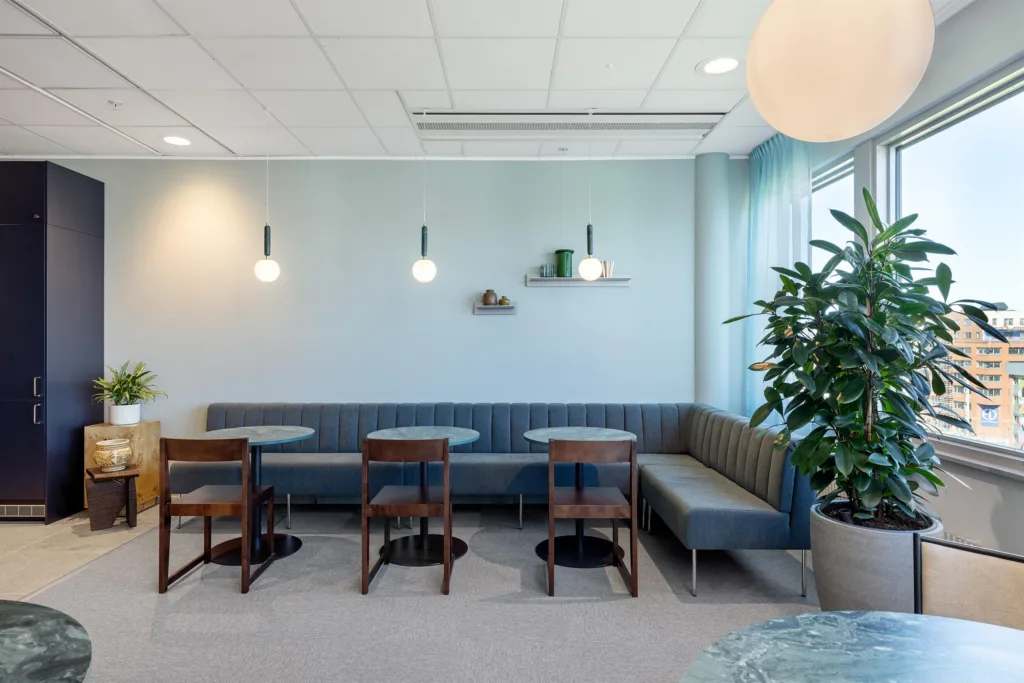 3. Ett ljust caféutrymme med blå soffbänkar, trästolar, små runda bord och vägglampor samt en växt vid fönstret.