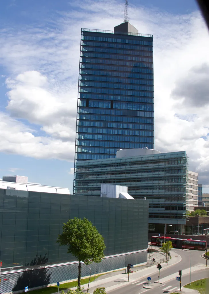En modern skyskrapa av glas med flera våningar under en molnig himmel, omgiven av andra byggnader.