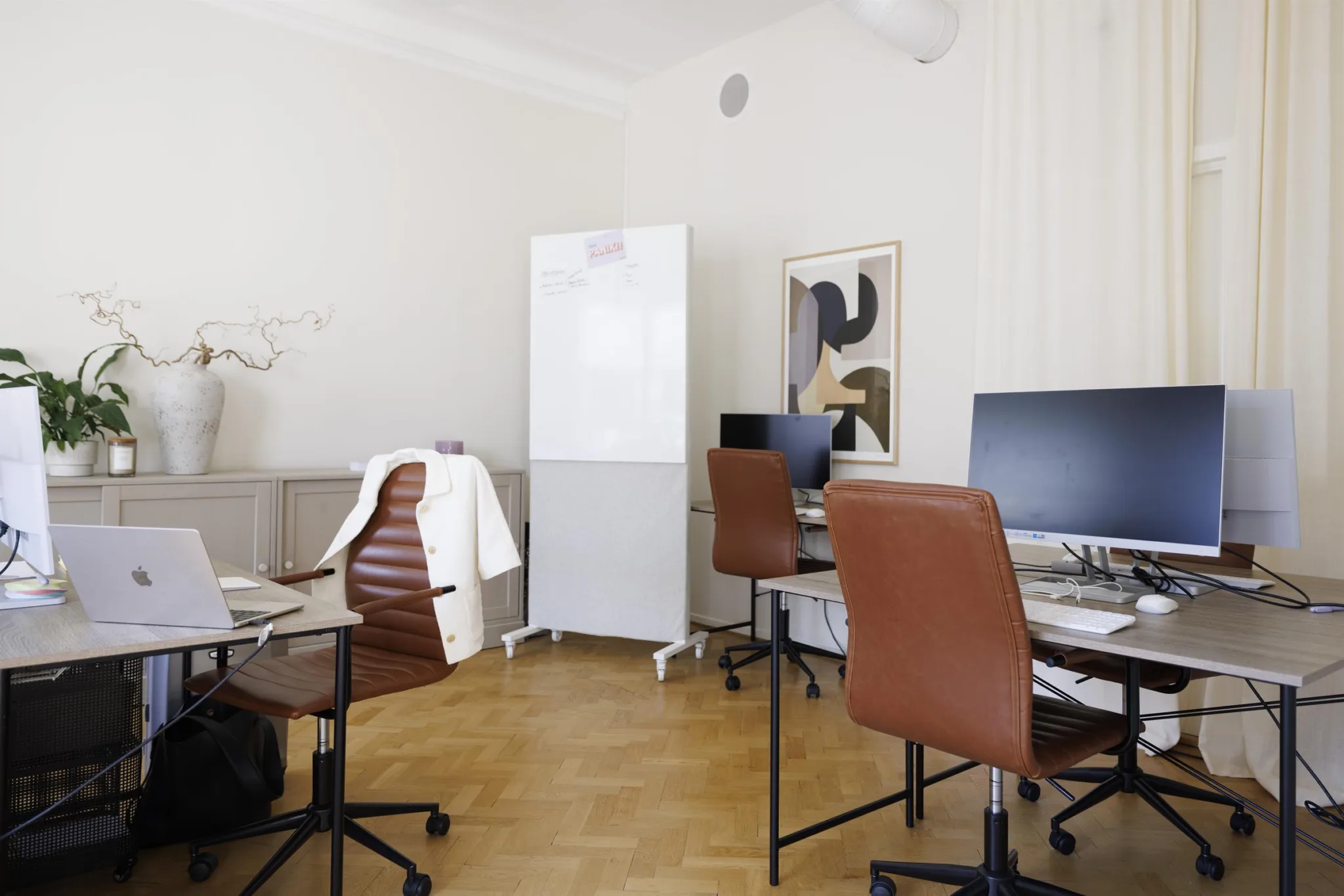 3. Ett kontorsrum med skrivbord, datorer, bruna stolar och en vit förvaringsskåp, dekorerat med en tavla på väggen.