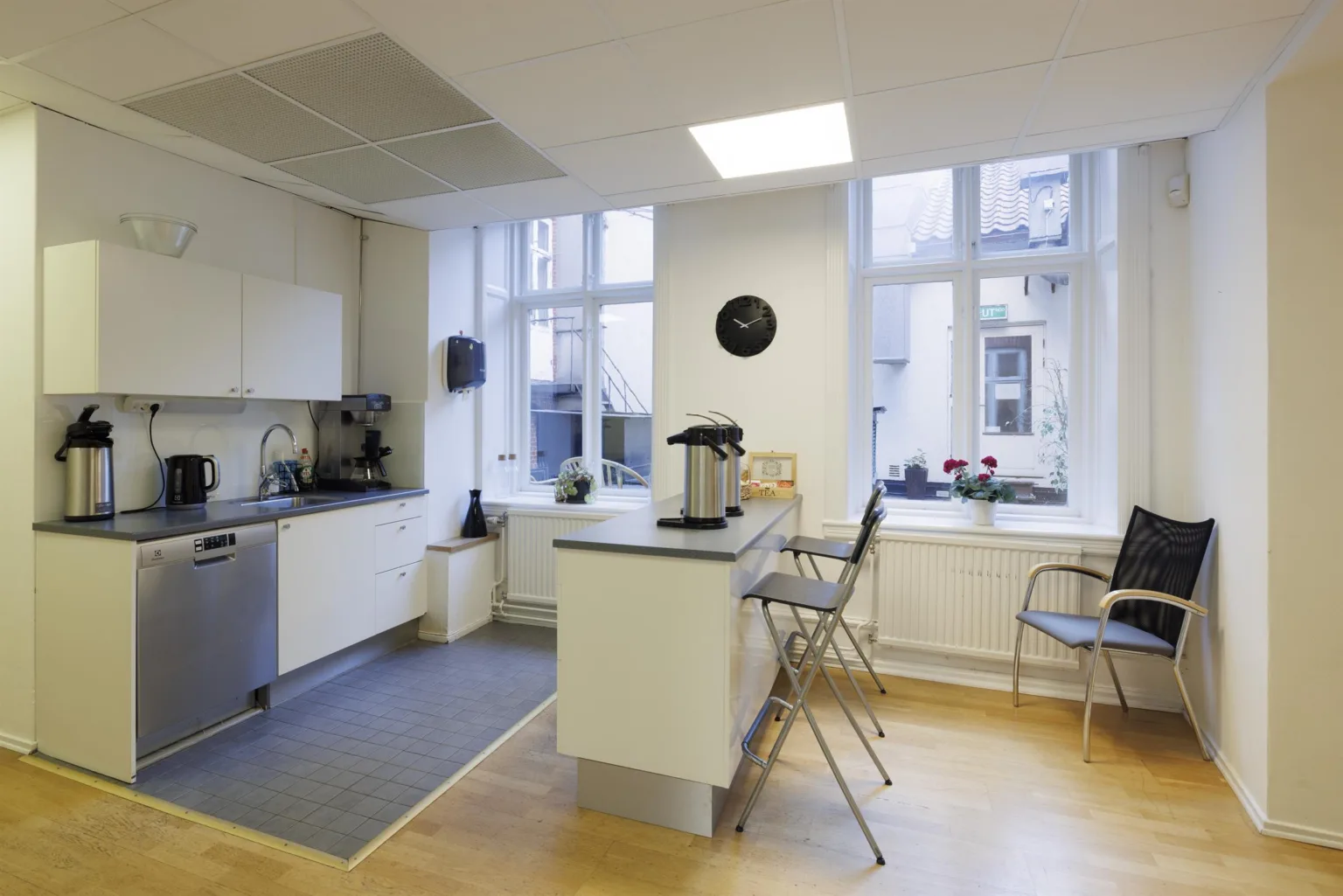 Ett modernt kök med vita skåp, en köksö med två barstolar och stora fönster som vetter mot en innergård.