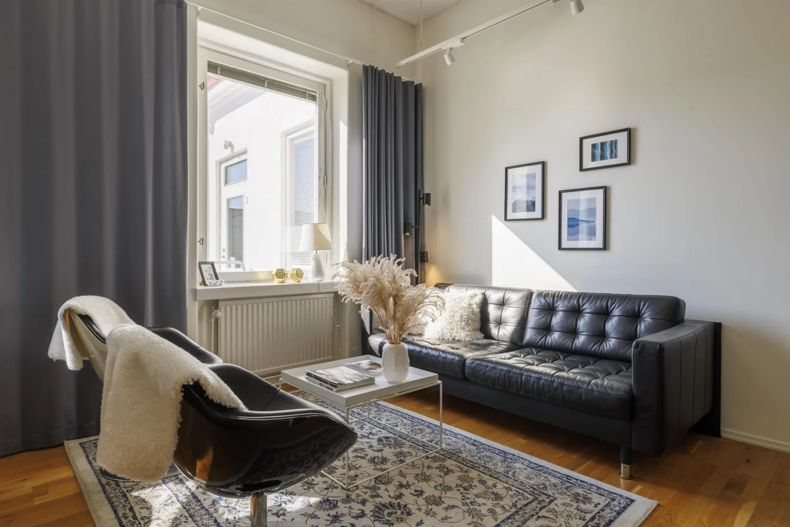 5. Ett vardagsrum med svart soffa, fåtölj, ljus matta och väggdekorationer bredvid ett stort fönster.