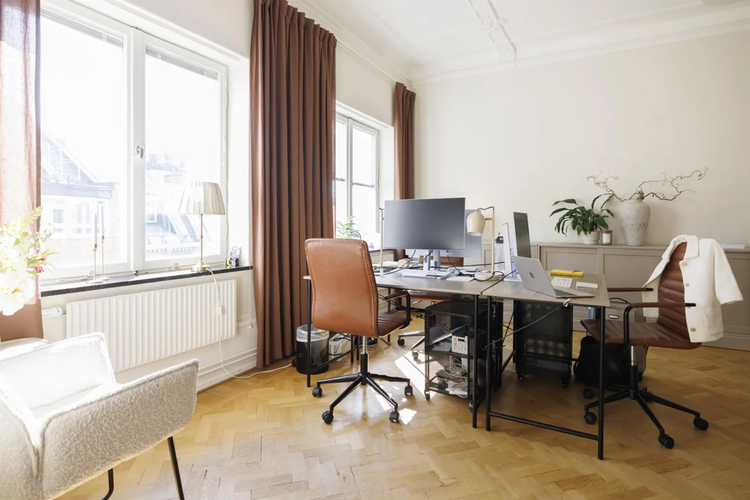 2. Ett hemmakontor med skrivbord, kontorsstol och dator framför stora fönster med bruna gardiner.