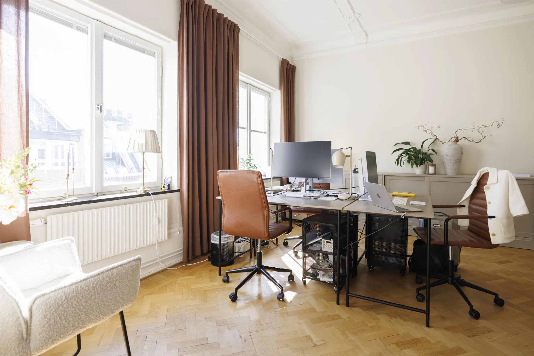2. Ett hemmakontor med skrivbord, kontorsstol och dator framför stora fönster med bruna gardiner.