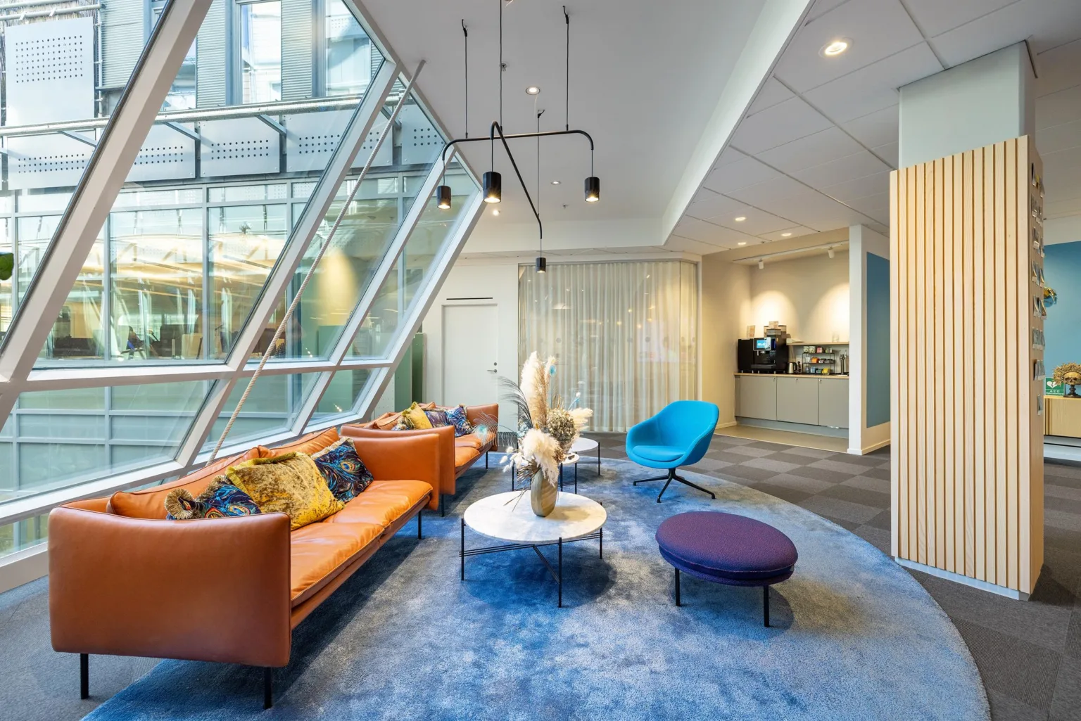 Ett modernt loungeområde med orange soffa, blå fåtölj och bord på en tjock blå matta, bredvid stora glasfönster.