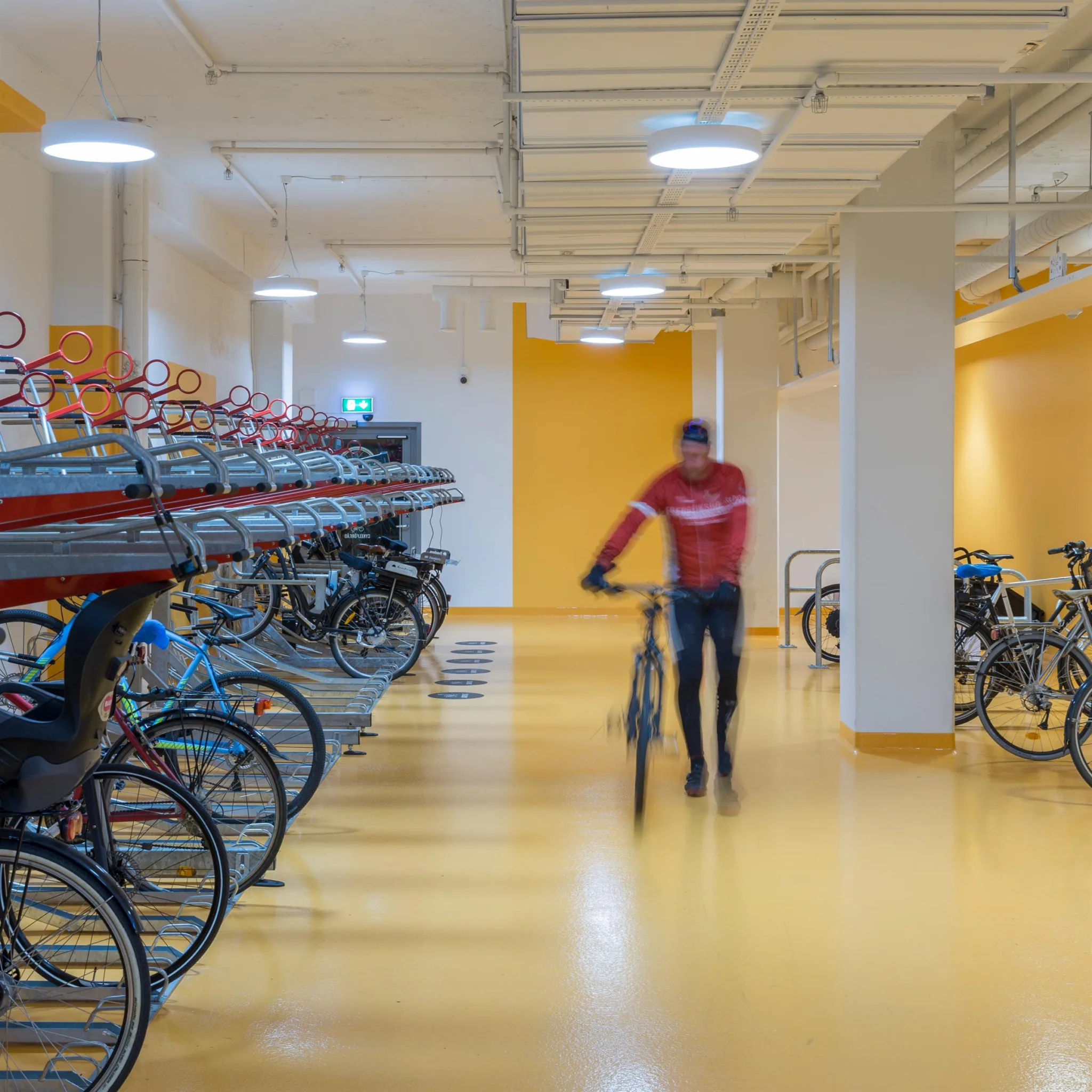 En cykelparkering inomhus med flera våningar av ställ, en person går med en cykel i en ljus lokal med gul golv.