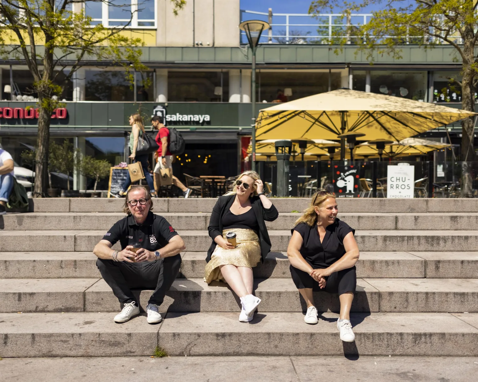 Tre personer sitter på trappsteg utomhus framför ett café med gula parasoller.