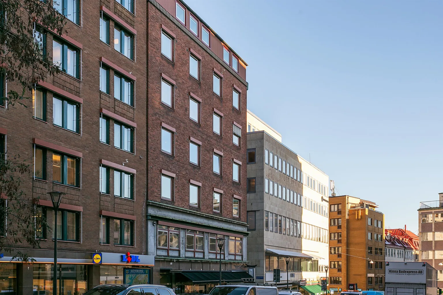 En gata med flervåningshus i olika arkitektonisk stil, parkerade bilar och klar blå himmel.