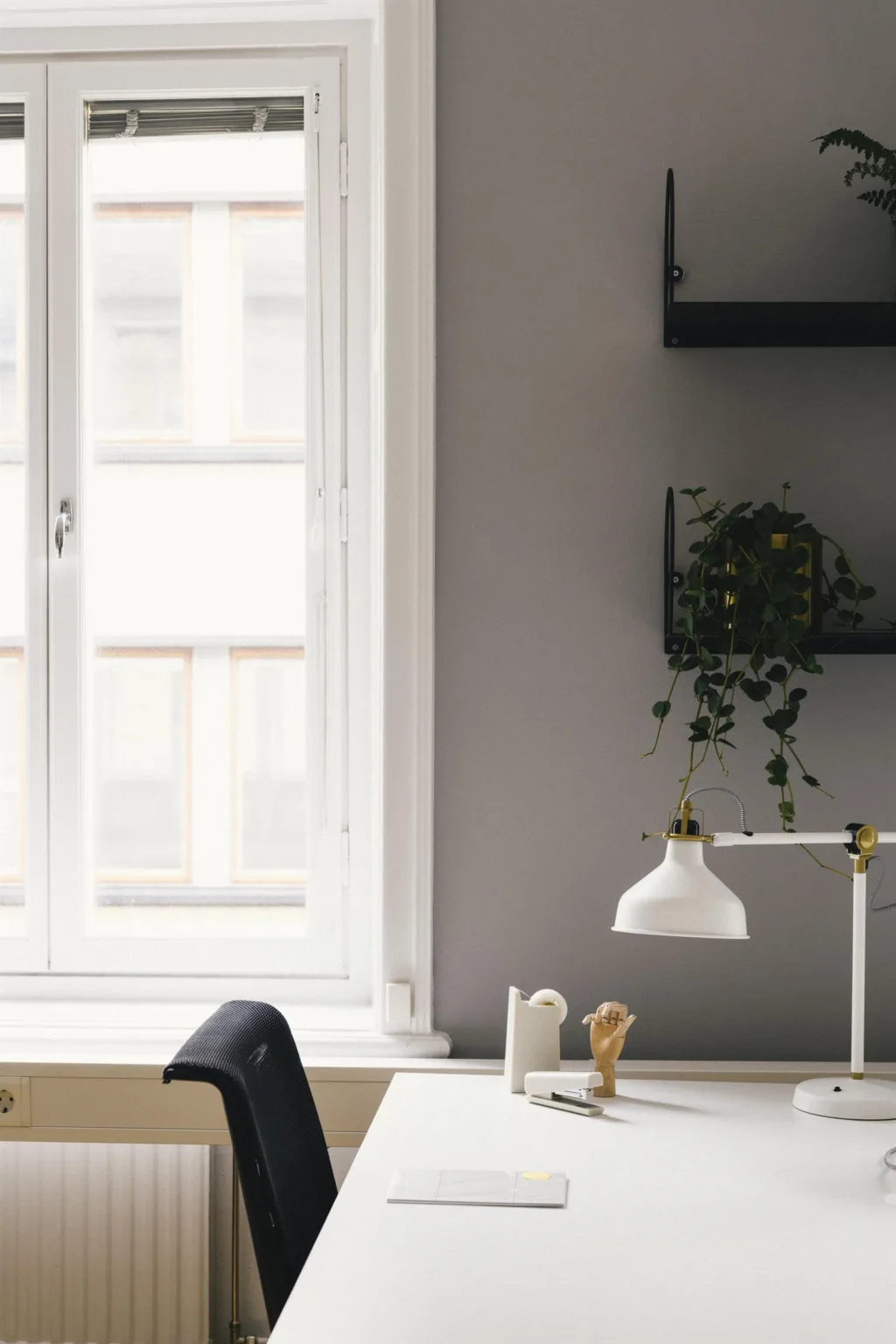 Ett skrivbord vid ett fönster med en vit lampa, dekorationer och en vägg med hyllor och en hängande växt.