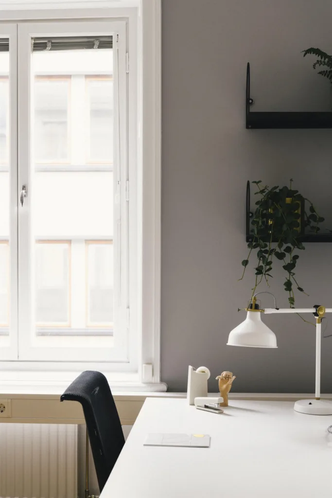 Ett skrivbord vid ett fönster med en vit lampa, dekorationer och en vägg med hyllor och en hängande växt.