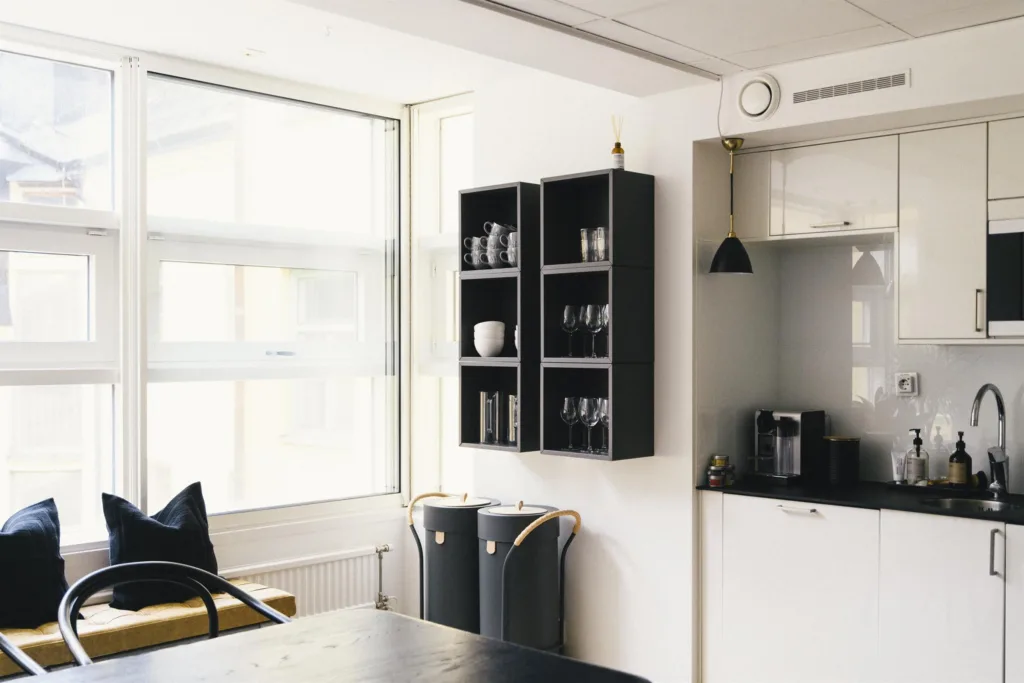 Ett kök med svarta väggmonterade hyllor, två svarta behållare och en kaffemaskin bredvid ett stort fönster.