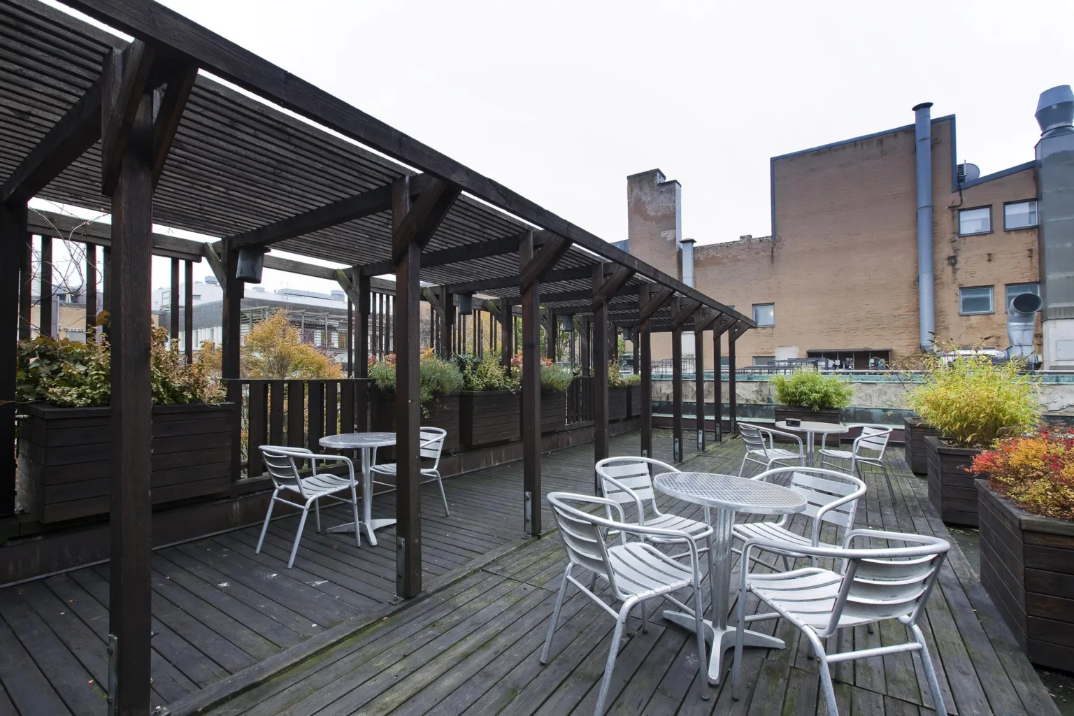 En utomhusterrass med trämöbler, pergola och växter, omgiven av byggnader.