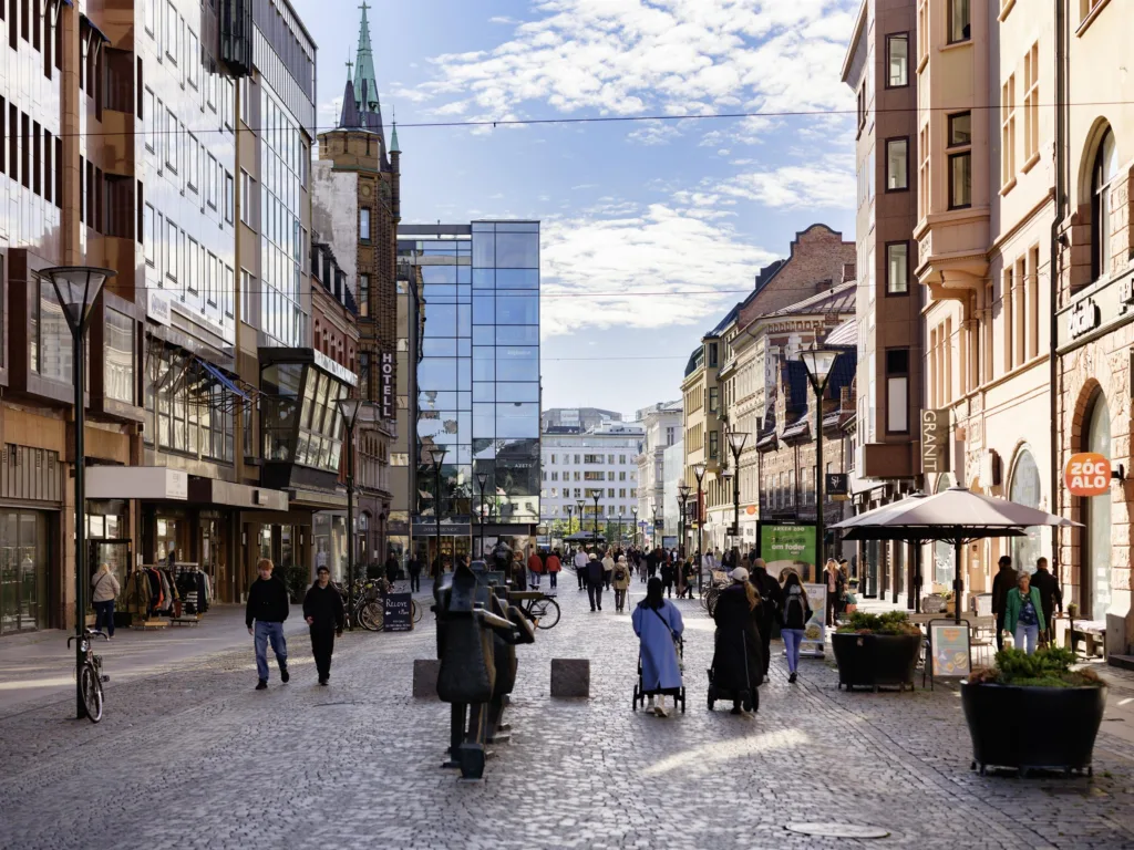 En livlig stadsgata med människor som går, omgivna av butiker, kaféer och höga byggnader; himlen är delvis molnig.