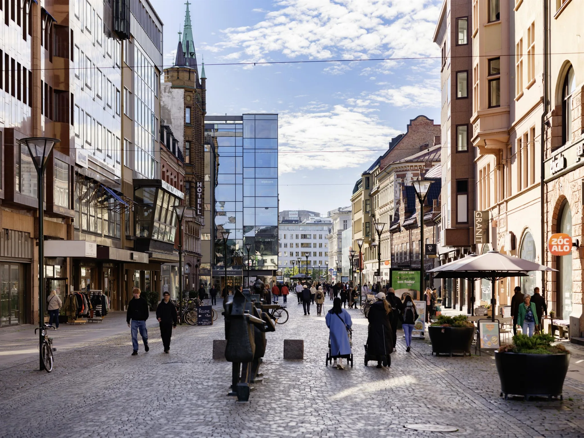 En livlig stadsgata med människor som går, omgivna av butiker, kaféer och höga byggnader; himlen är delvis molnig.