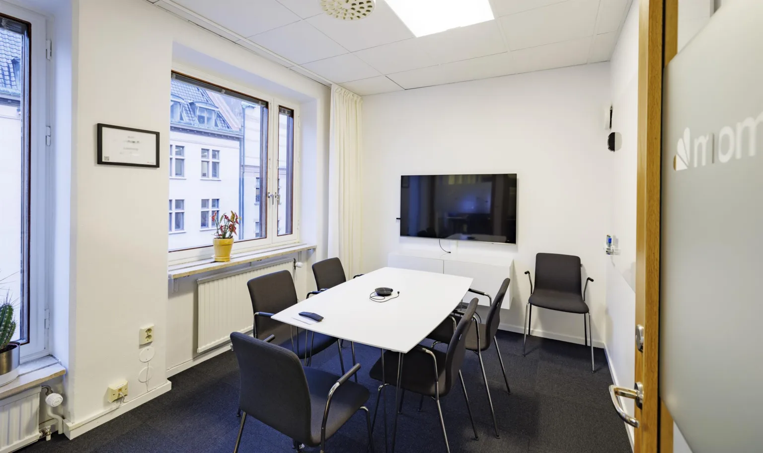 Ett litet konferensrum med ett bord, sex stolar och en väggmonterad TV; ett fönster med utsikt över byggnader syns till vänster.