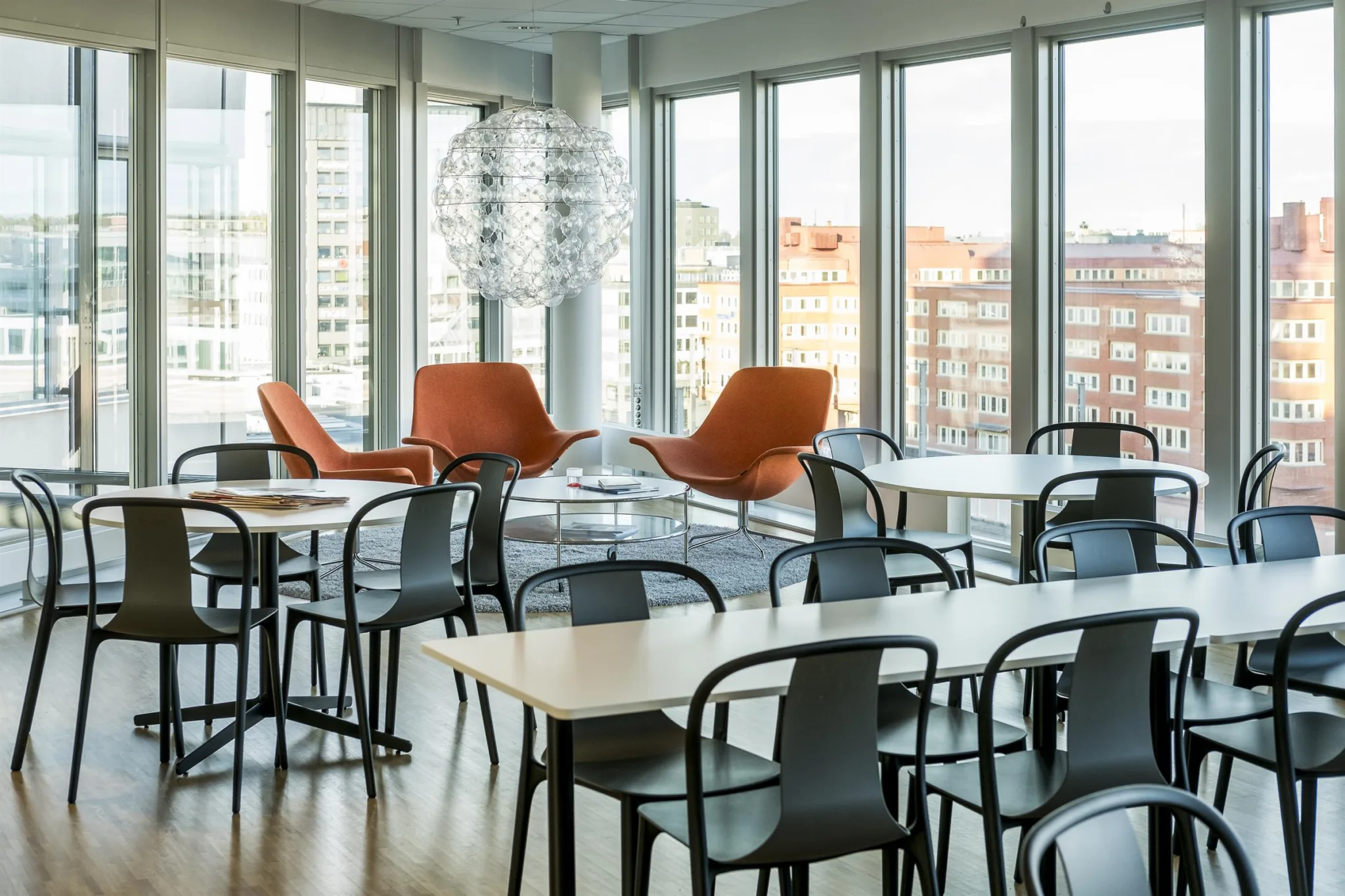 Ett modernt lunchrum med bord, svarta stolar, orange fåtöljer och stora fönster med stadsutsikt.
