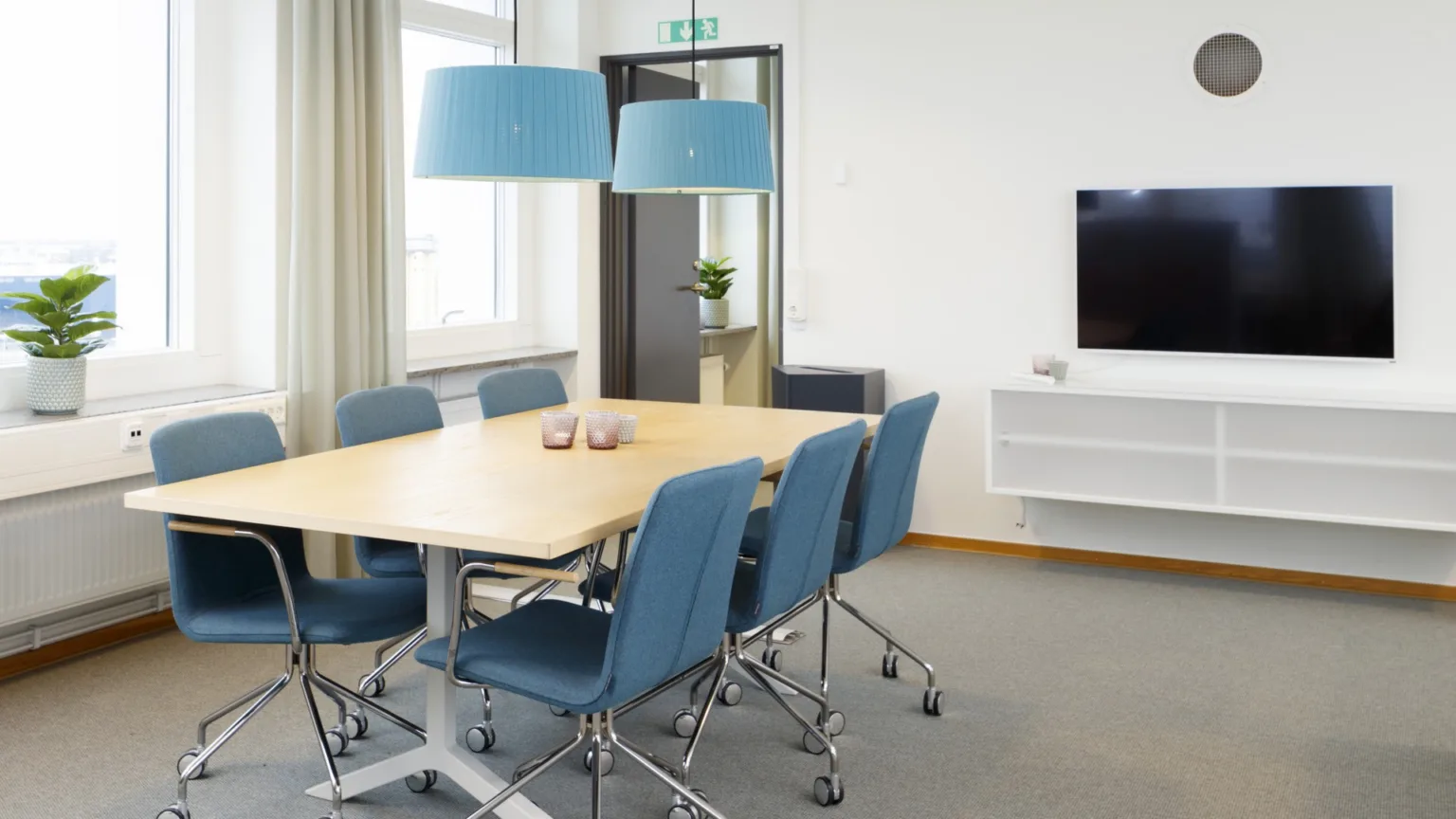 3. Mötesrum med träbord, blå stolar, två blå taklampor och en väggmonterad TV.