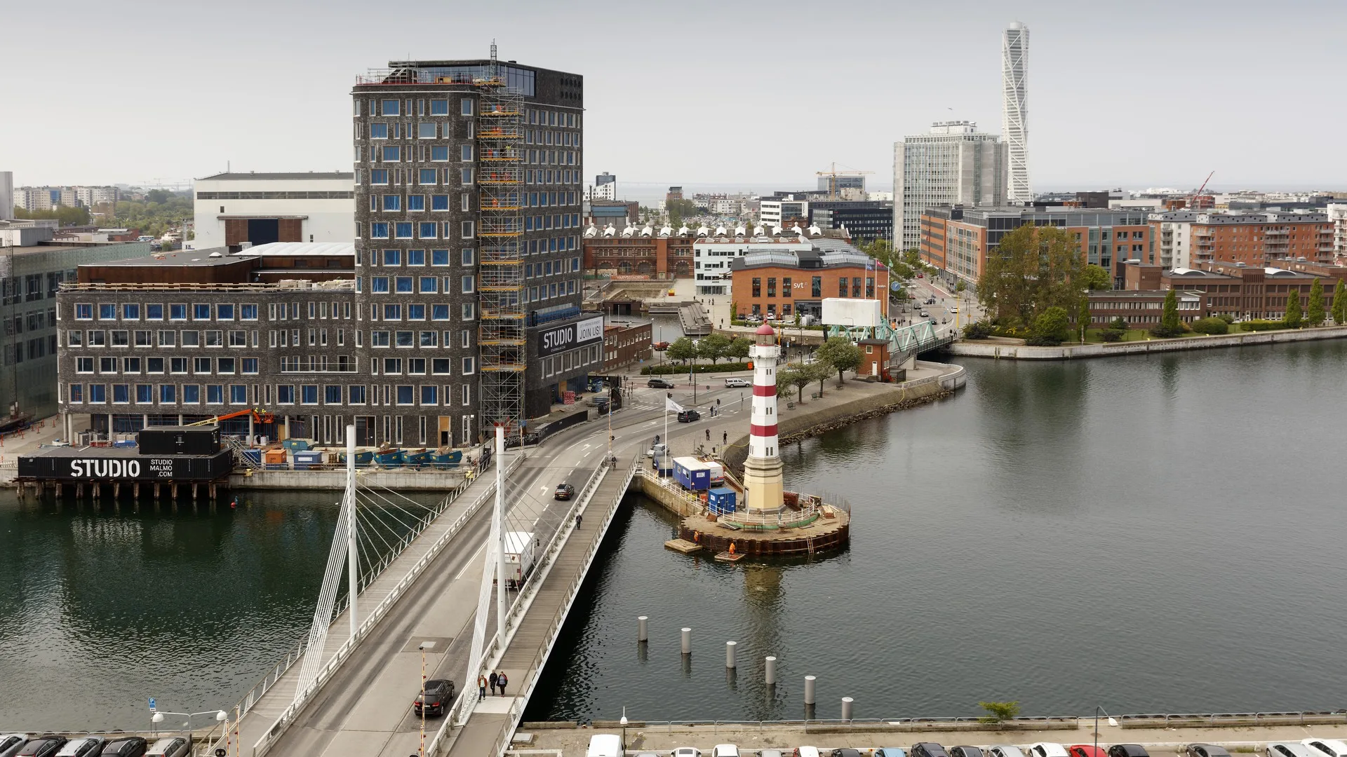 2. En stadsvy med en bro, en fyr omgiven av vatten och moderna byggnader i bakgrunden.