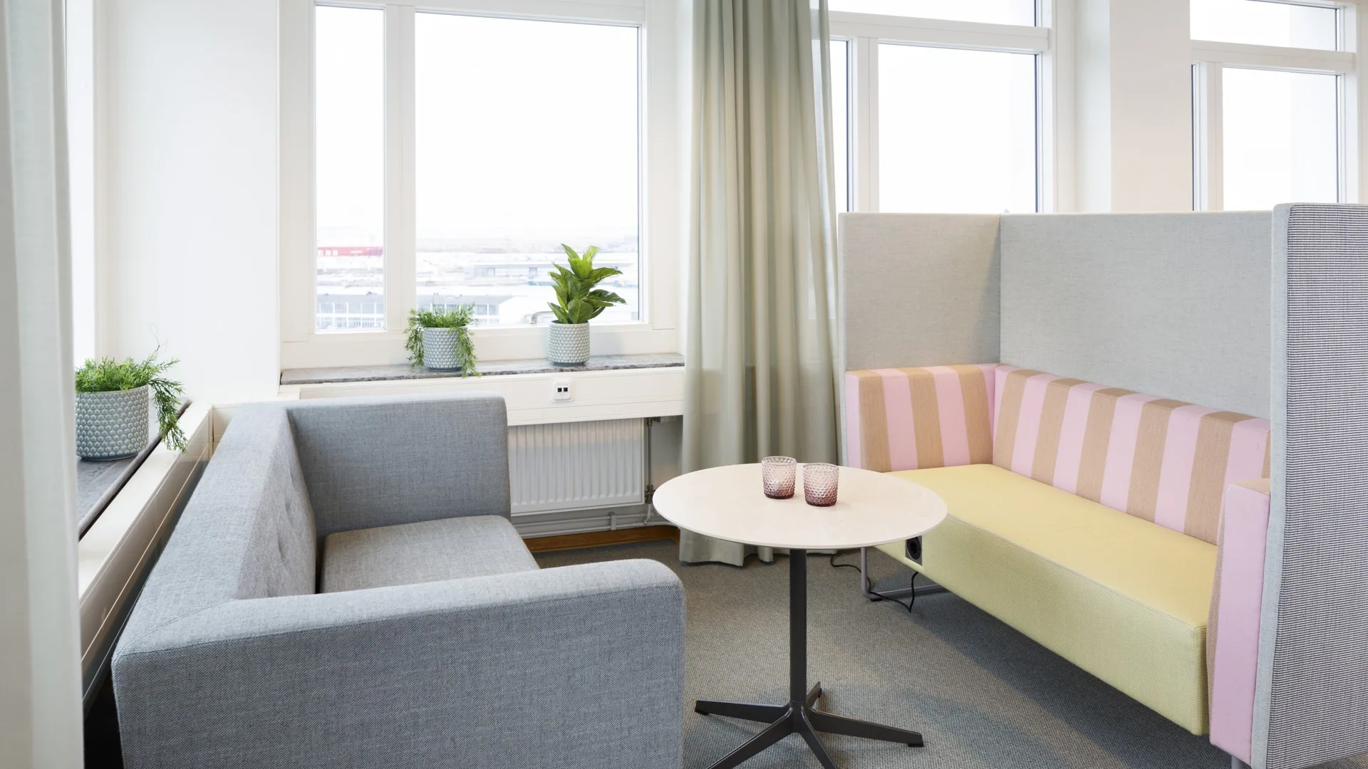 Ett litet vardagsrum med en grå soffa, en gul- och rosarandig fåtölj, ett runt bord och fönster med gardiner.