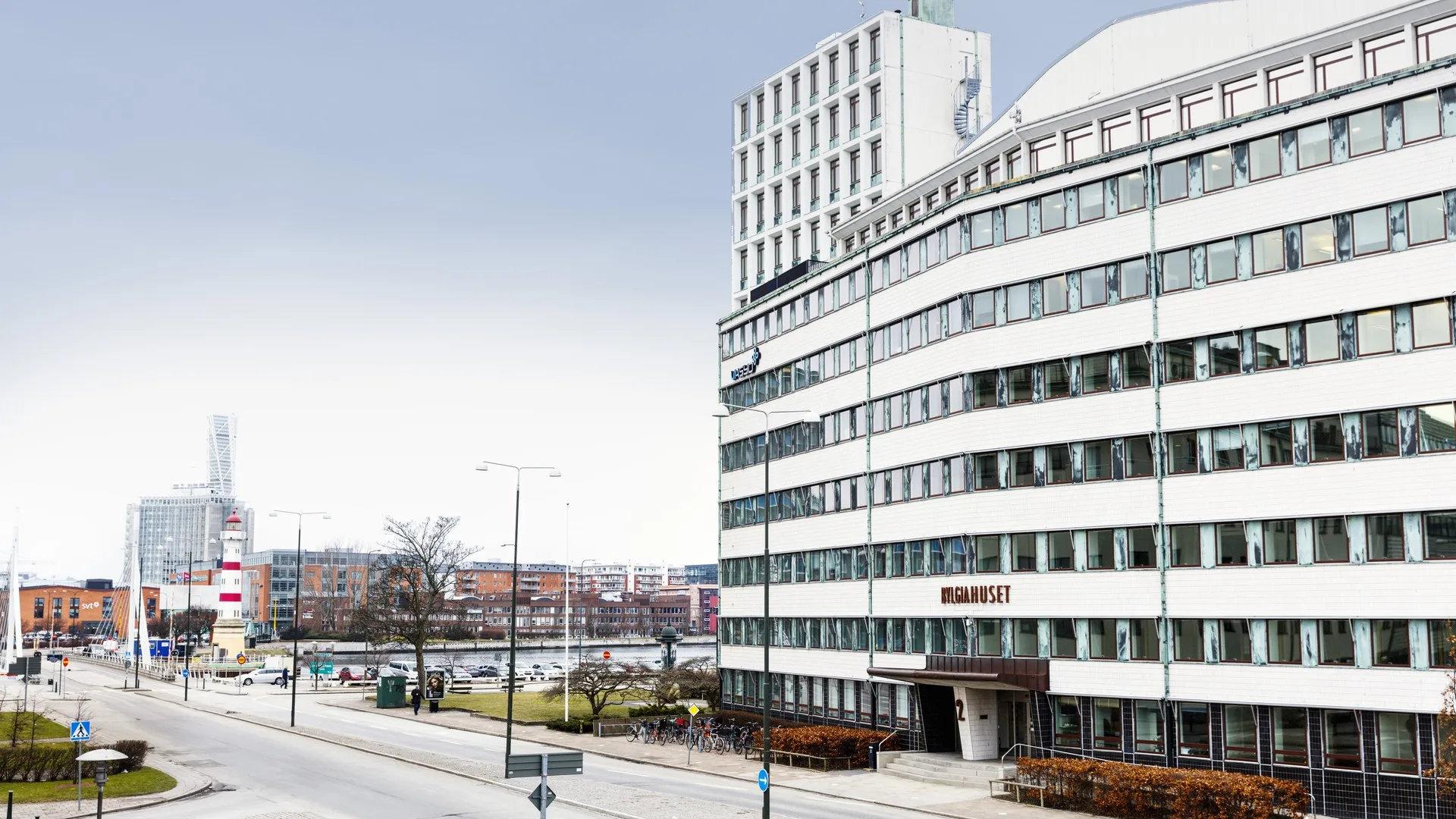 1. En vit kontorsbyggnad med flera våningar och stora glasfönster, belägen vid en gata med utsikt över en kanal och omgivande stadsmiljö.