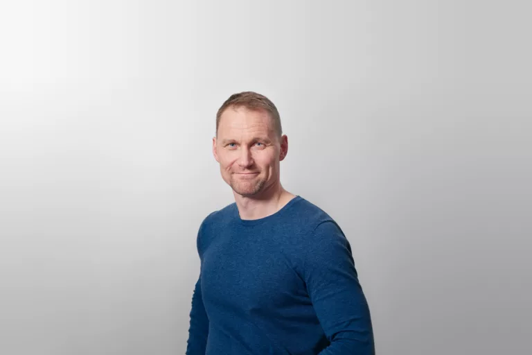 En person med kort hår, klädd i en blå tröja, står mot en ljusgrå bakgrund.