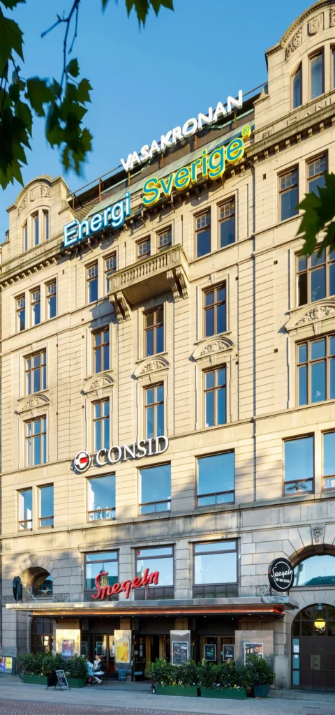 En beige byggnad med skyltarna "Energi", "Consid" och andra företagsskyltar. Fönstren reflekterar ljuset och trädkronor syns delvis.