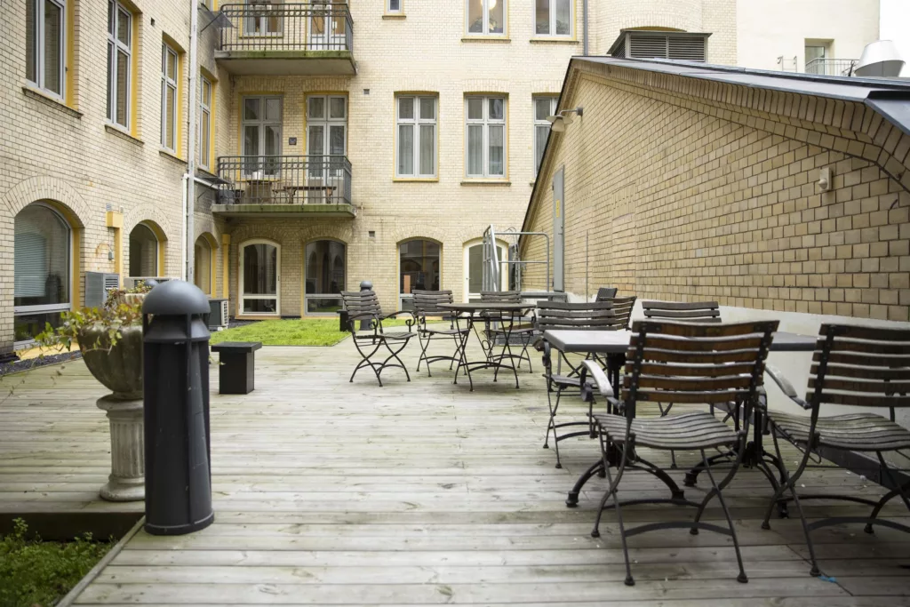 En innergård med trätrall, bord och stolar omgivna av beige byggnader med stora fönster.