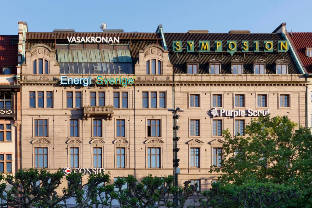 En stor byggnad med skyltar som "Vasakronan", "Energi", och "Symposion", omgiven av träd och blå himmel.