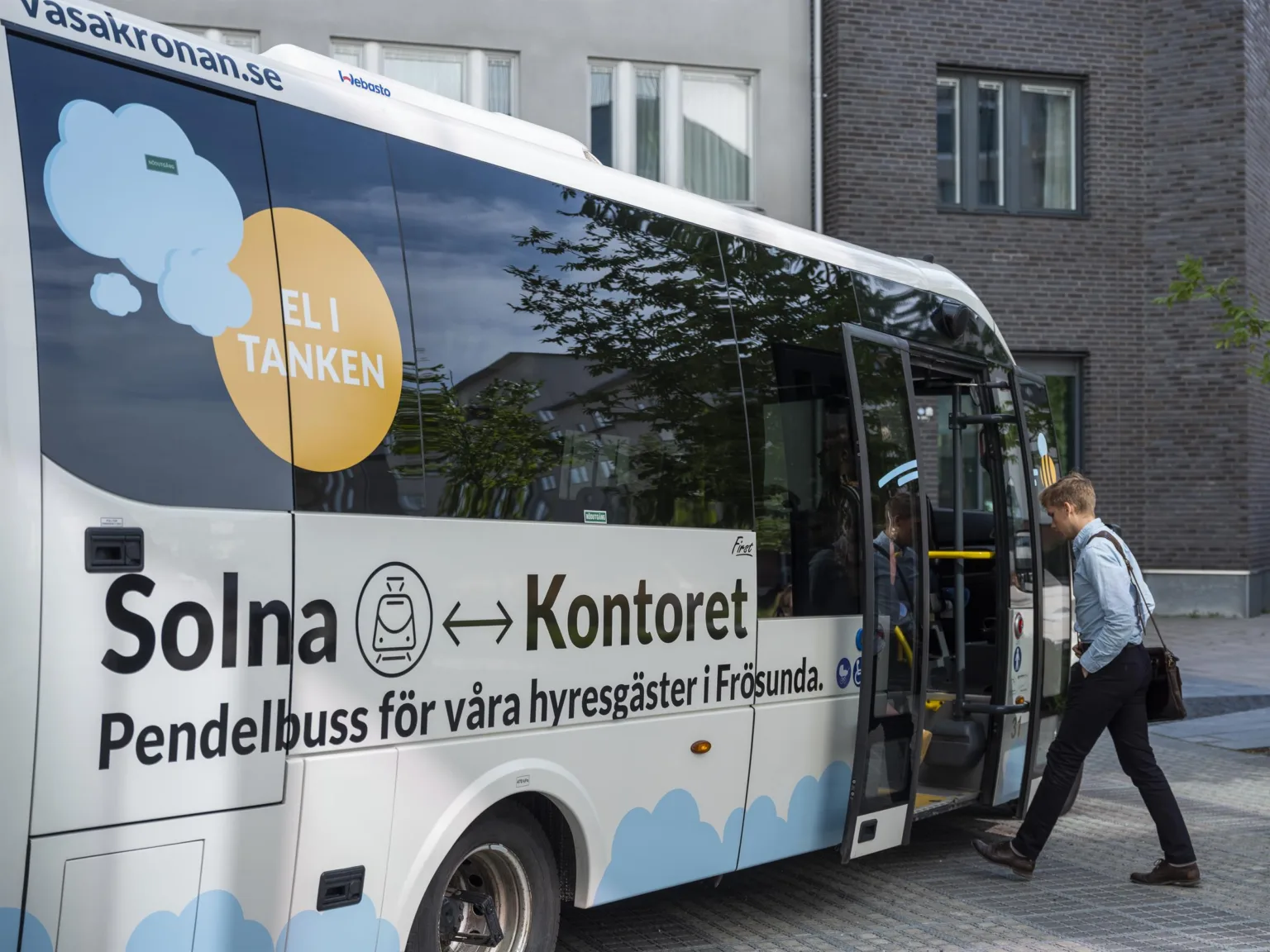 En vit eldriven pendelbuss med texten "Solna ↔ Kontoret" och "Eldriven pendelbuss för våra hyresgäster i Frösunda," parkerad vid en trottoar medan en person går ombord.