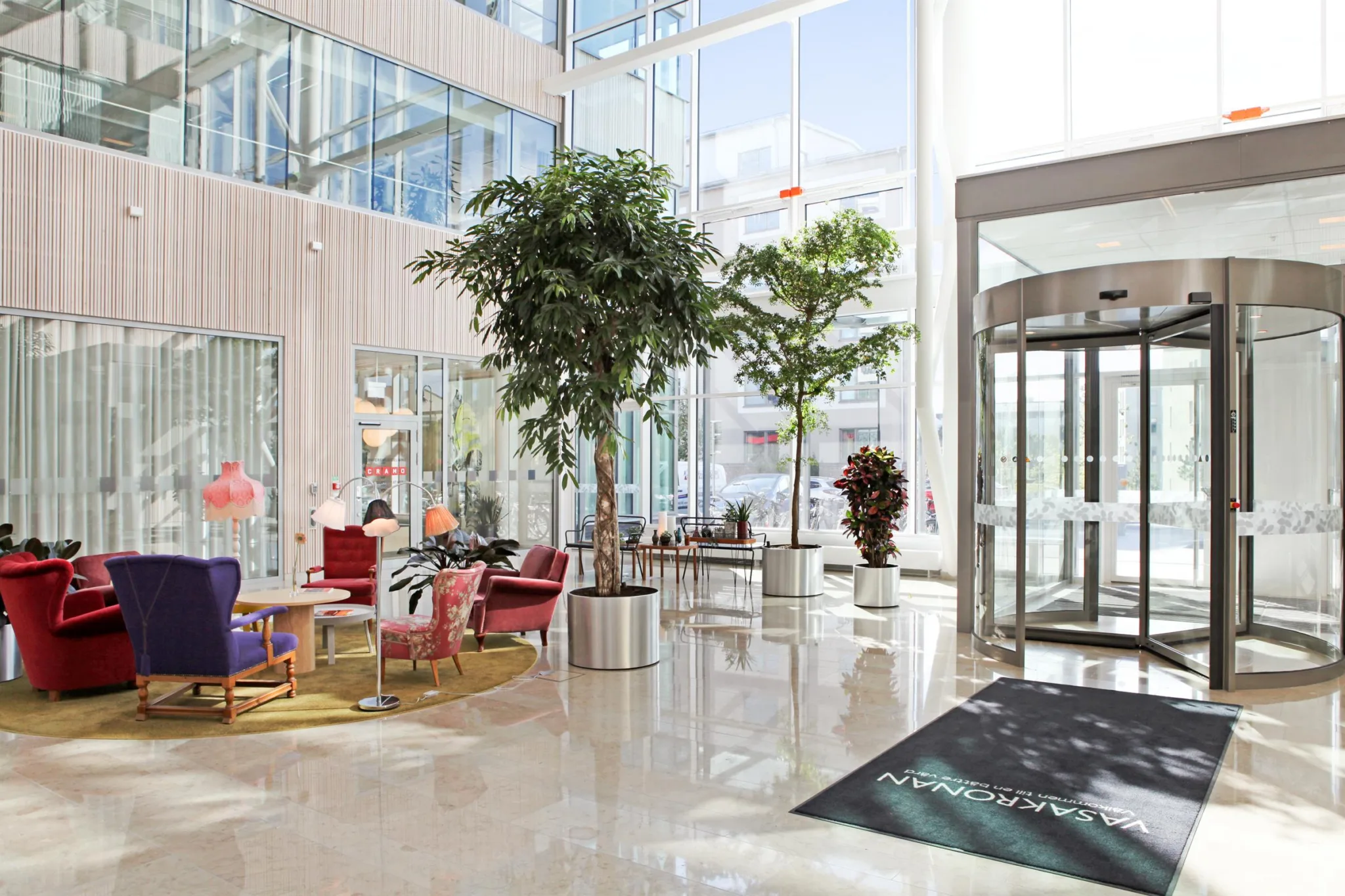 En ljus lobby med stora fönster, växter i krukor, färgglada möbler och en roterande entrédörr.