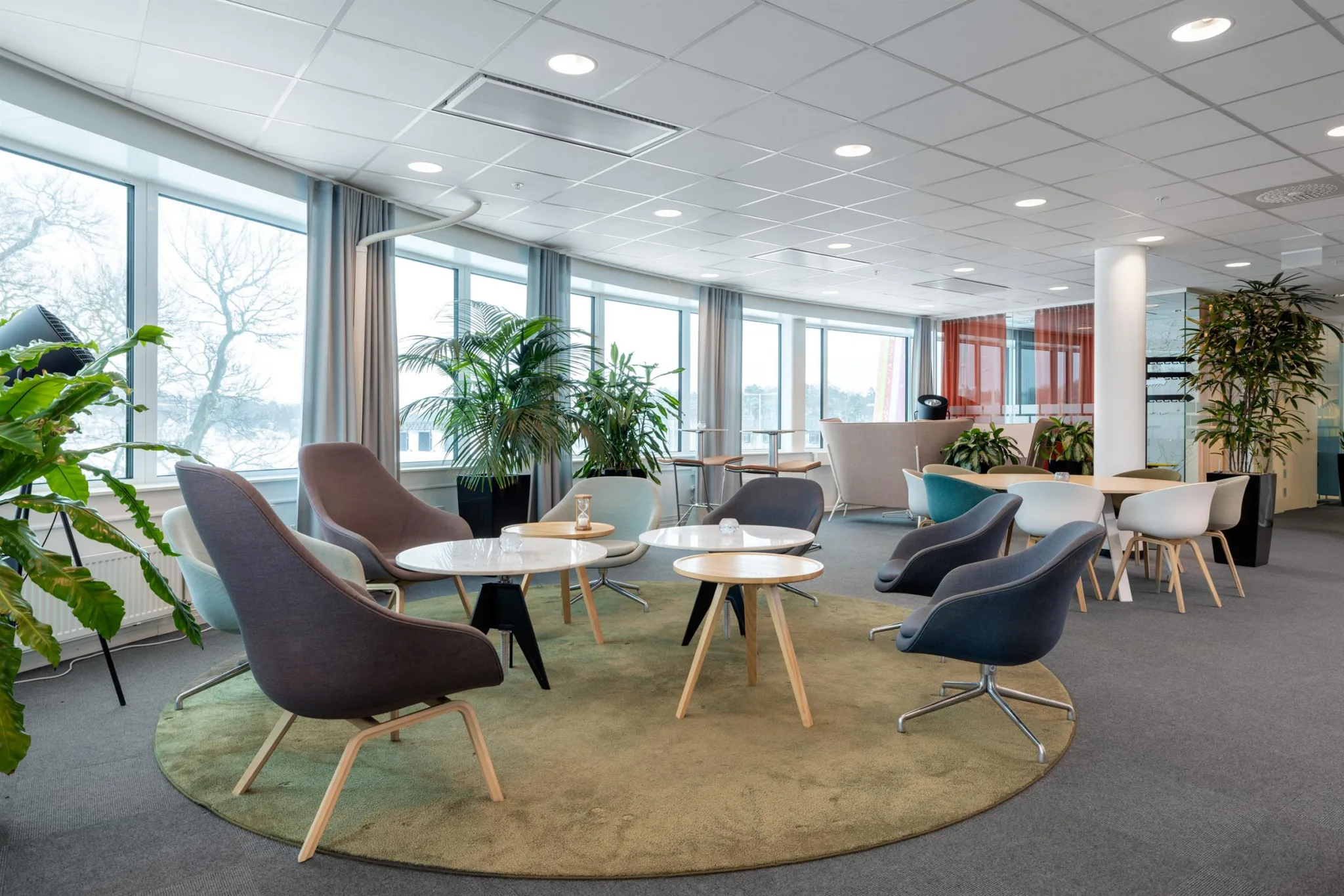 3. Loungeområde med bekväma stolar, små bord, gröna växter och stora fönster som ger utsikt.