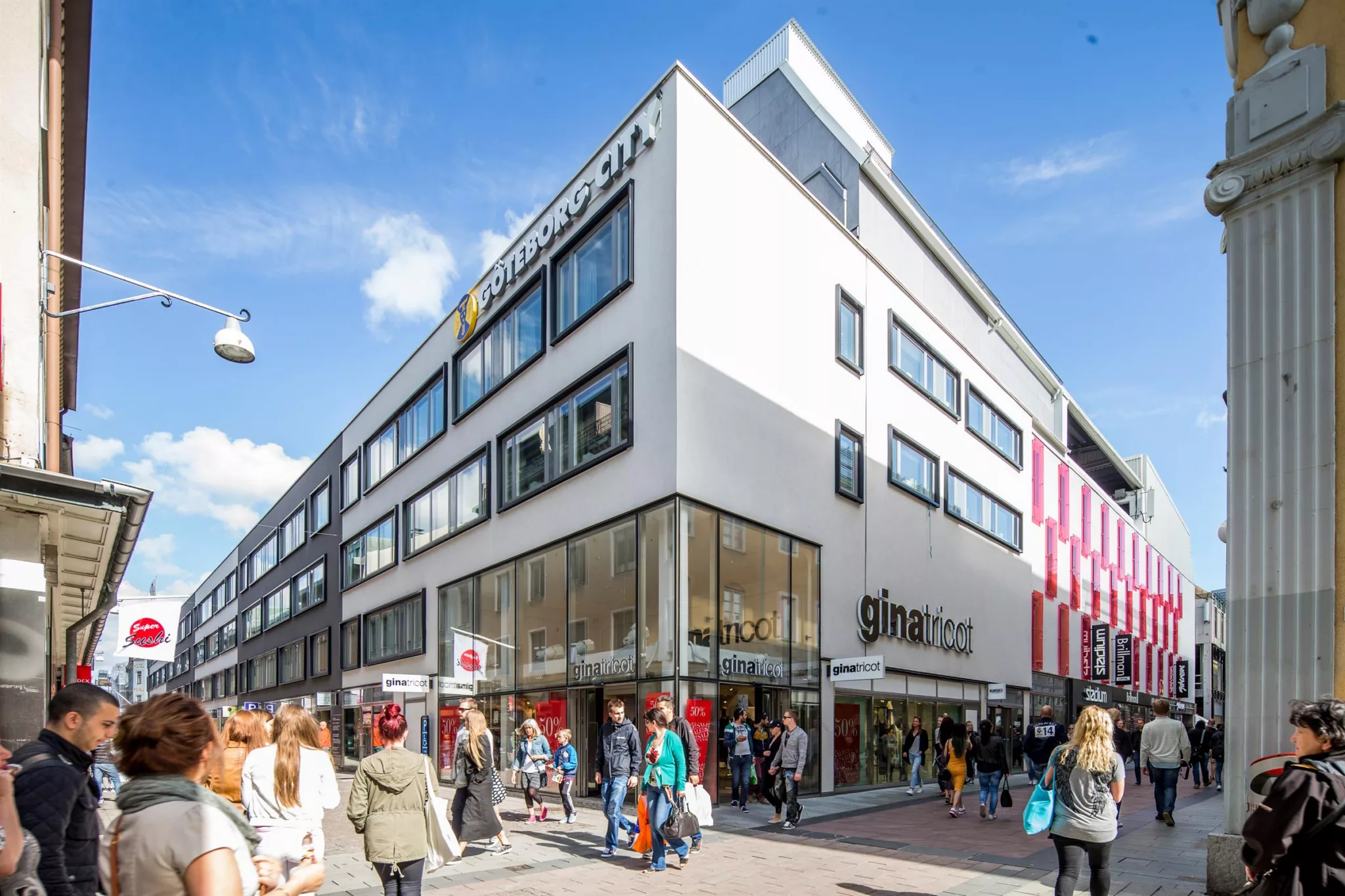 En vit byggnad med stora fönster och skyltar för Galleria Center Syd, omgiven av människor som promenerar under en blå himmel.