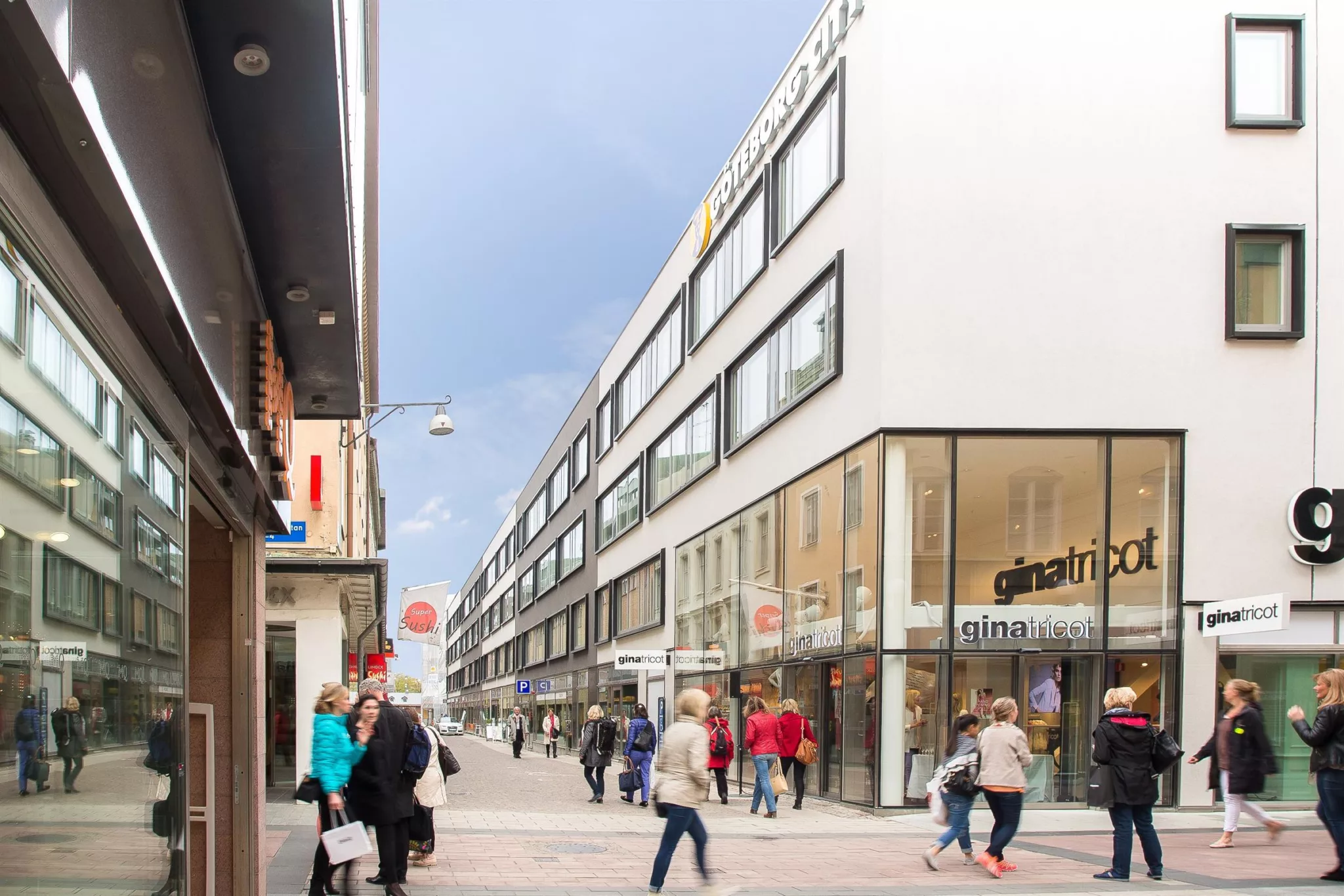 En stadsgata med människor som går förbi en vit byggnad med fönster och en skylt för Galleria Center Syd, under en molnig himmel.