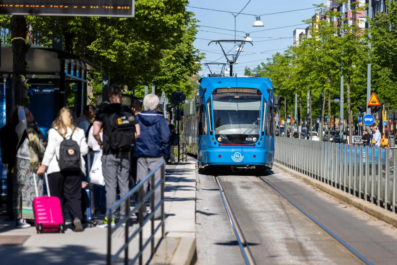 En blå spårvagn närmar sig en hållplats där människor väntar.