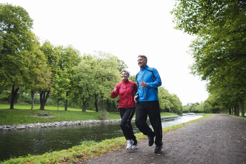 Två personer joggar bredvid en kanal, omgivna av grönskande träd och en stig.