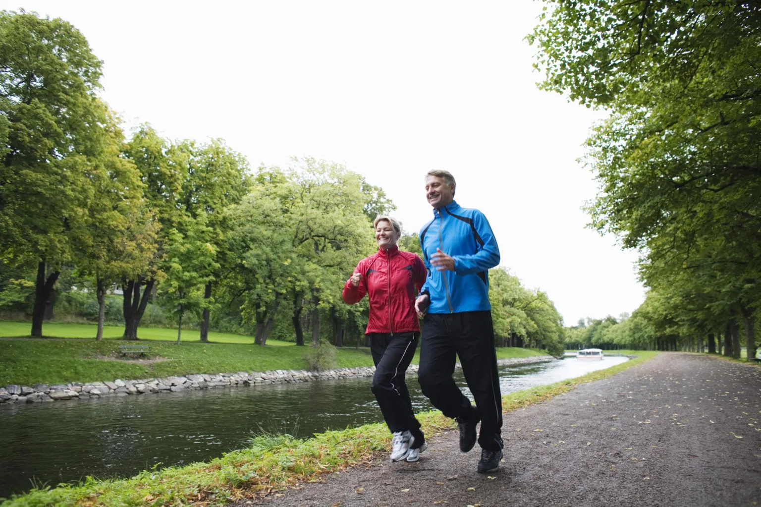 Två personer joggar bredvid en kanal, omgivna av grönskande träd och en stig.