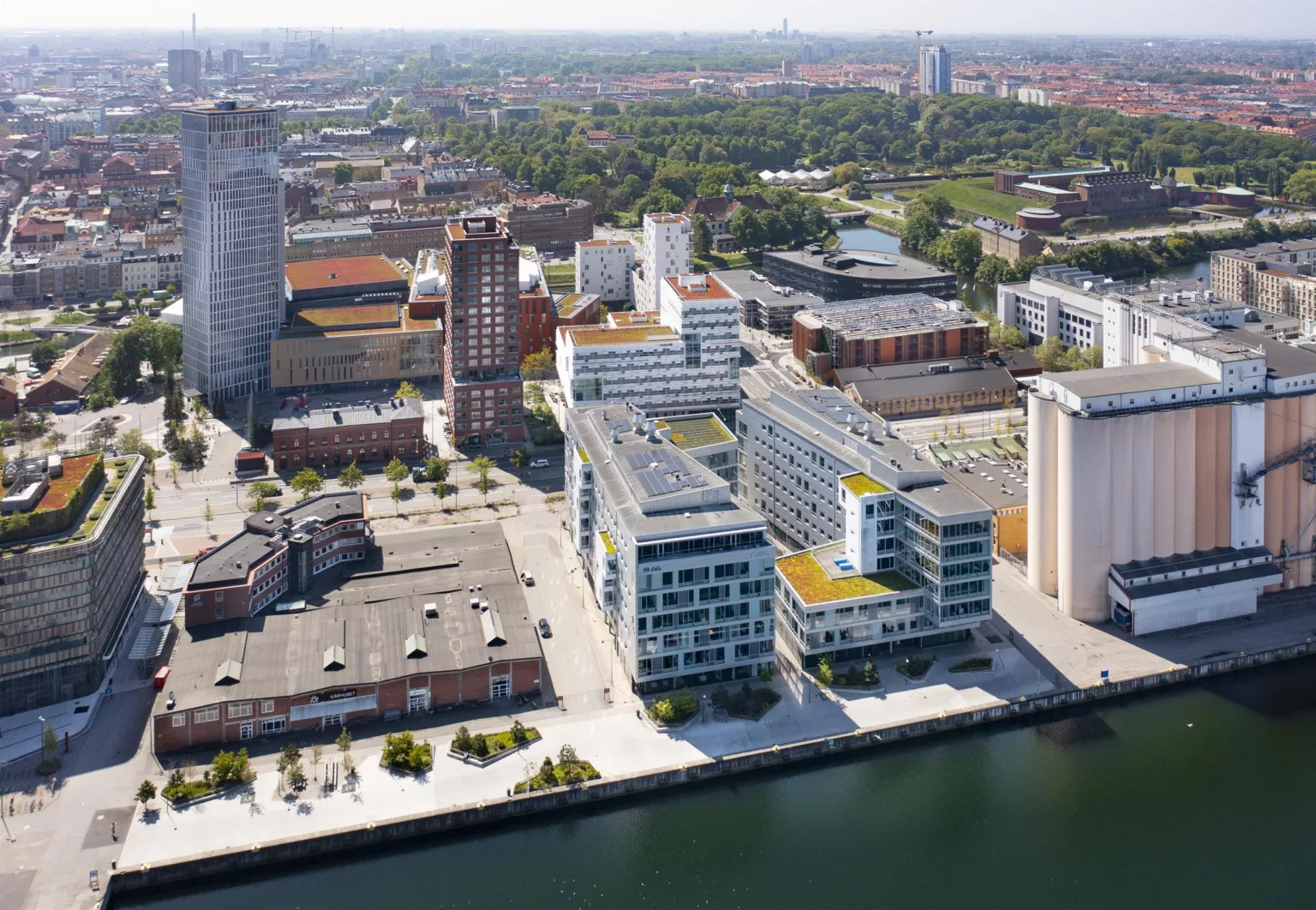 Flygfoto över en stadsdel med moderna byggnader, grönområden och en kanal i förgrunden.