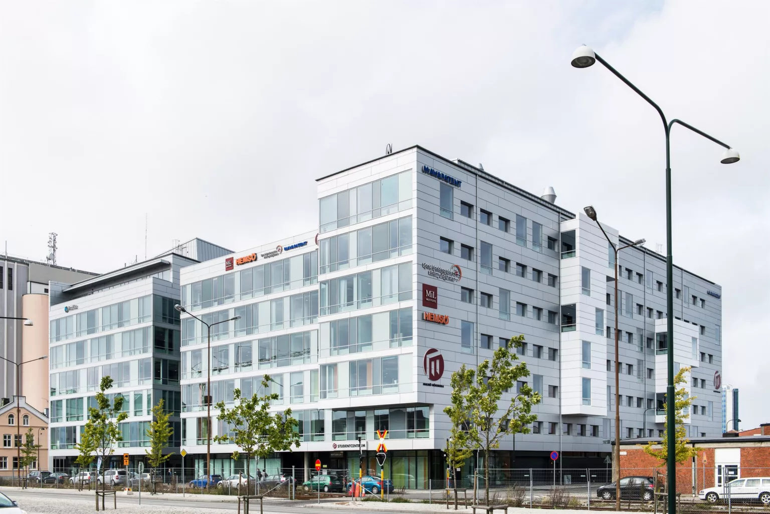 En vit modern byggnad med glasfasad och flera företagslogotyper, omgiven av träd och gatubelysning.