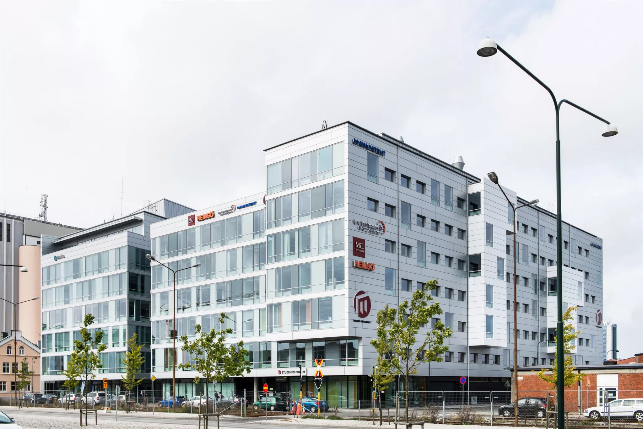 En vit modern byggnad med glasfasad och flera företagslogotyper, omgiven av träd och gatubelysning.