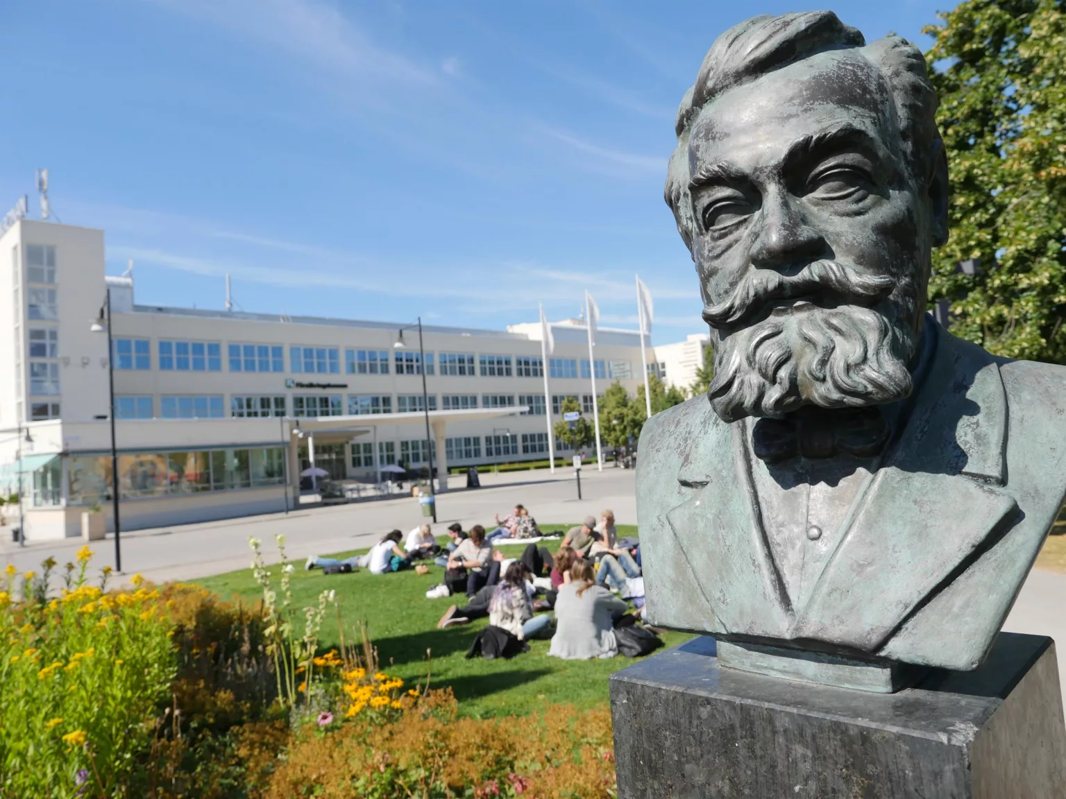 3. En staty av en man med skägg i förgrunden, med en grönskande gräsyta där människor sitter i bakgrunden. En modern byggnad syns längre bort.