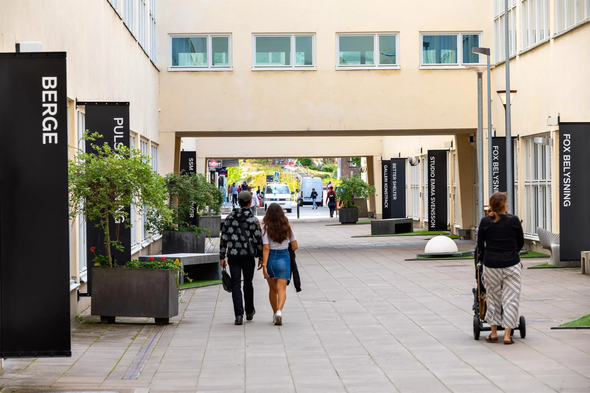4. Gångväg mellan byggnader där människor går, med gröna växter och bänkar längs kanterna. En ljus portal leder vidare ut mot ett öppet område.