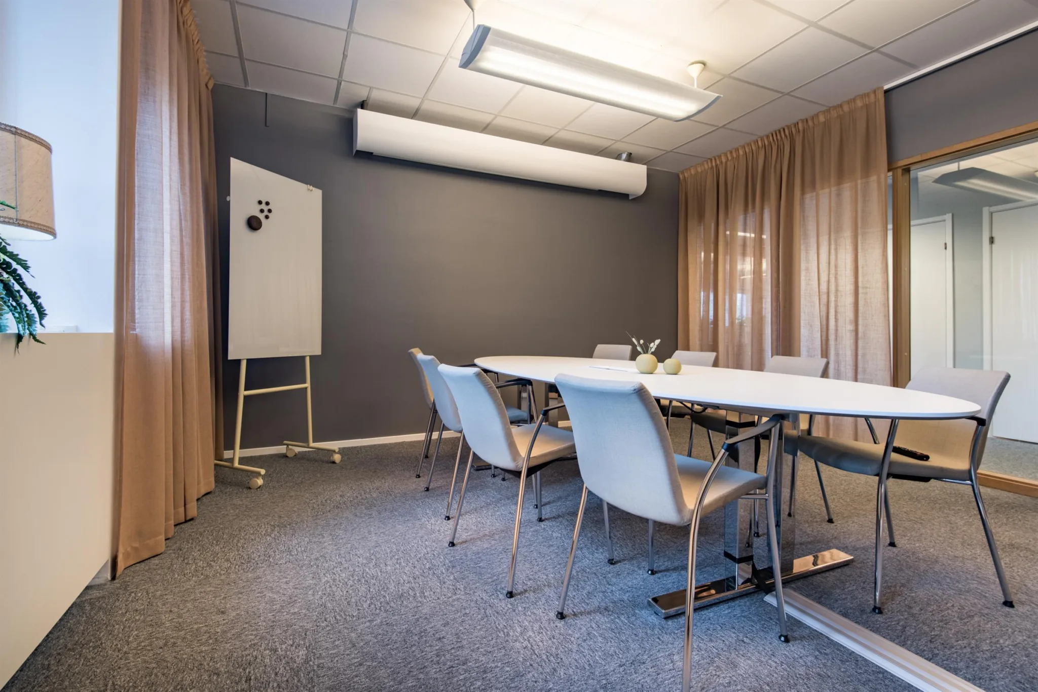 Ett konferensrum med vita stolar, ett långt bord, en whiteboard och beige gardiner.