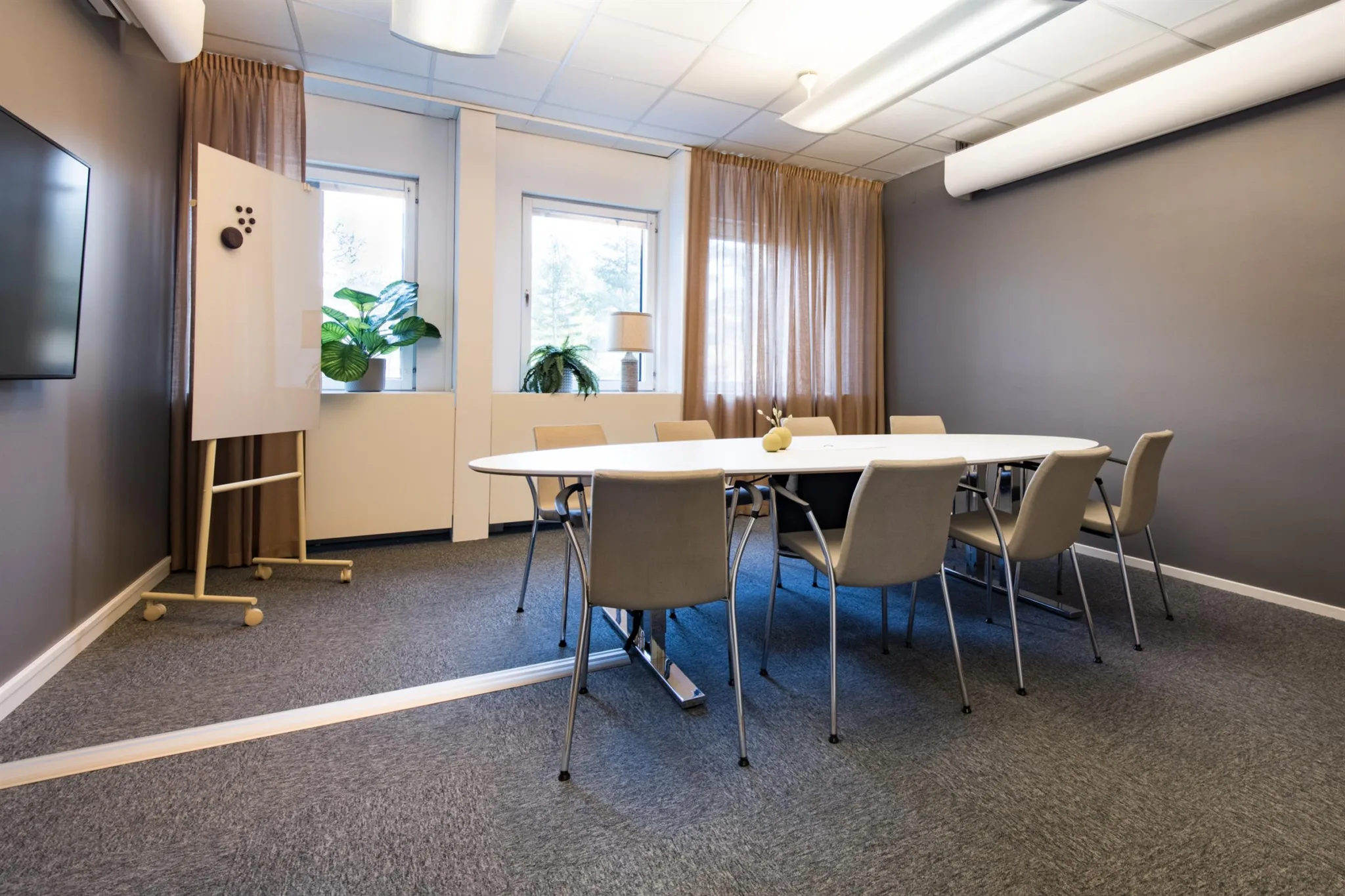 Ett mötesrum med ett ovalt bord, stolar, en whiteboard och stora fönster med gardiner som släpper in ljus.