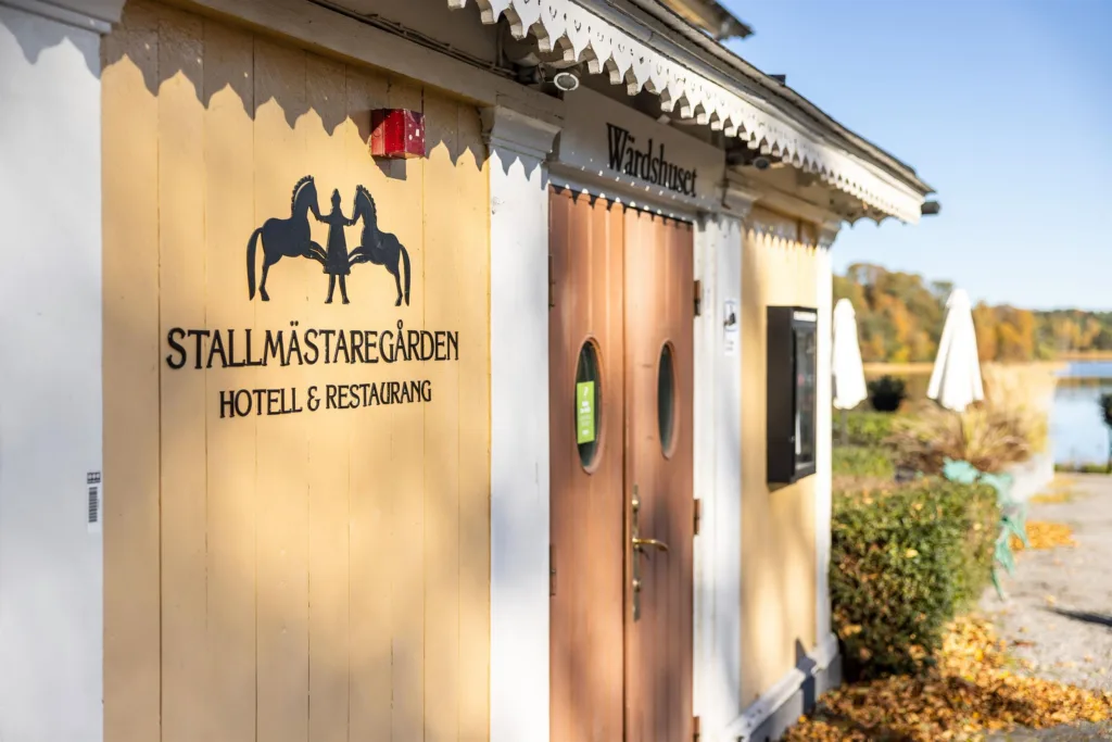 2. Gul byggnad med texten "TALLMÄSTAREGÅRDEN HOTELL & RESTAURANG" på väggen, en träport och omgivande grönska.