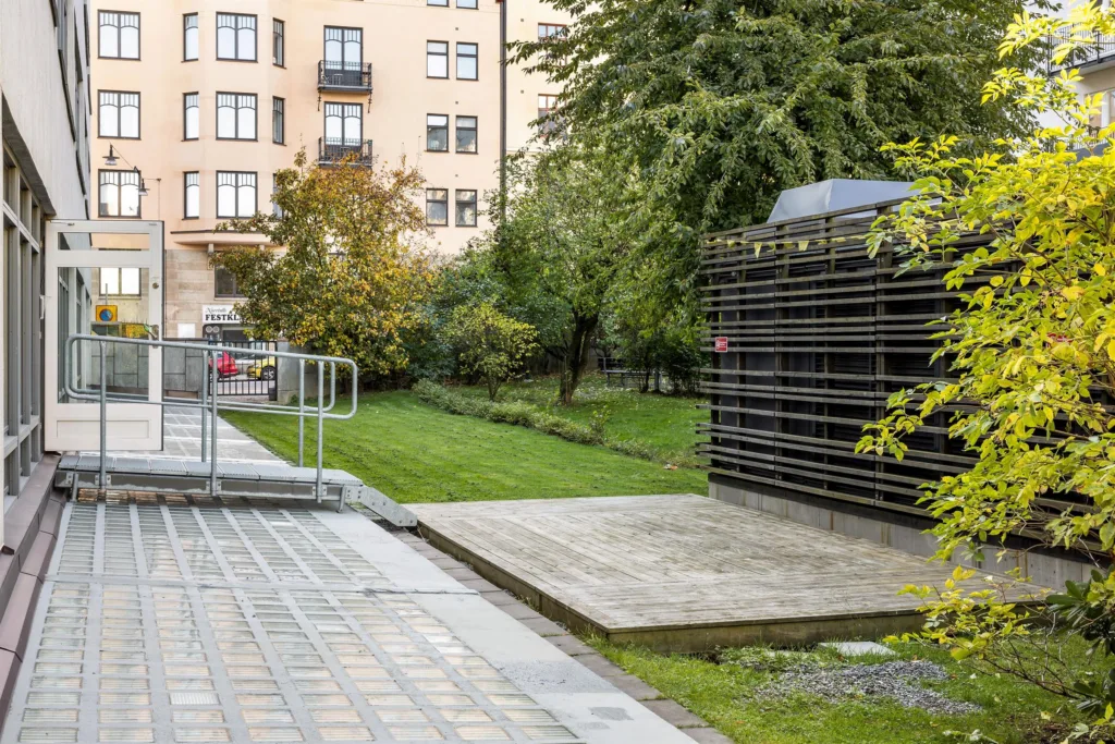 En utomhusmiljö med grönska, en byggnad i bakgrunden och en metallstruktur till höger.