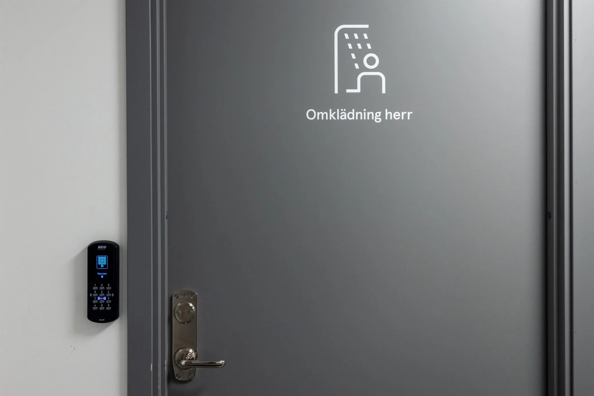 En grå dörr med en symbol som visar en dusch och texten "Omklädning herr" bredvid ett elektroniskt låssystem.