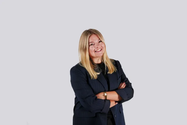 En kvinna med blont hår som ler, klädd i en mörk kavaj, mot en ljus bakgrund.