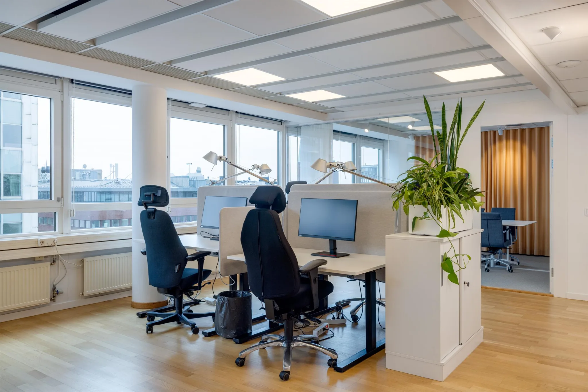 Öppen kontorsmiljö med skrivbord, datorskärmar och kontorsstolar, samt stora fönster som släpper in naturligt ljus.