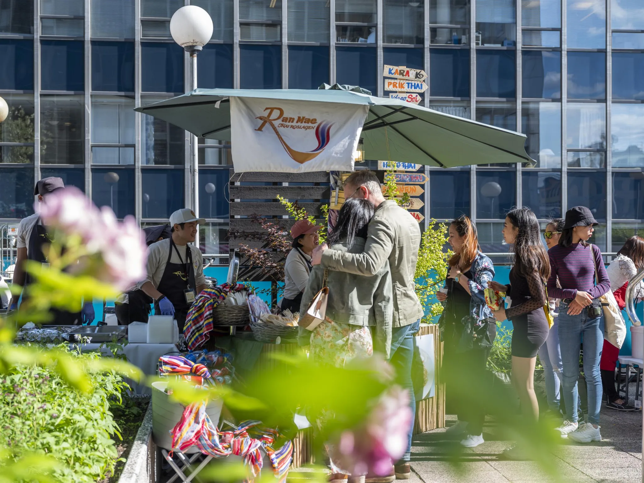 En utomhusmarknad med människor som handlar vid ett stånd med grönsaker och blommor, omgiven av stadens höga byggnader.