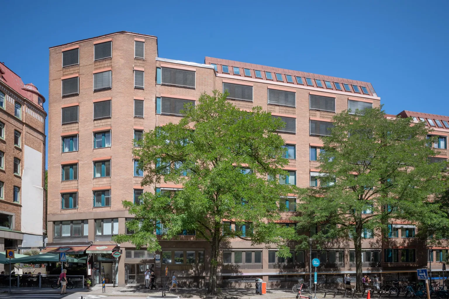 En röd tegelbyggnad med stora fönster, omgiven av grönskande träd och en trottoar med människor.